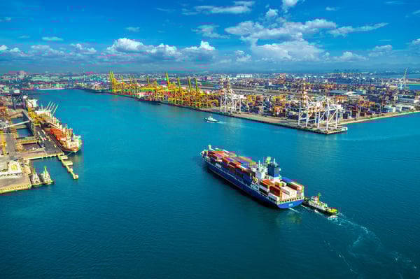 A cargo ship navigation filled with colored containers navigating to a port, in the blue sea, with blue sky and clouds, representing what exporting means.